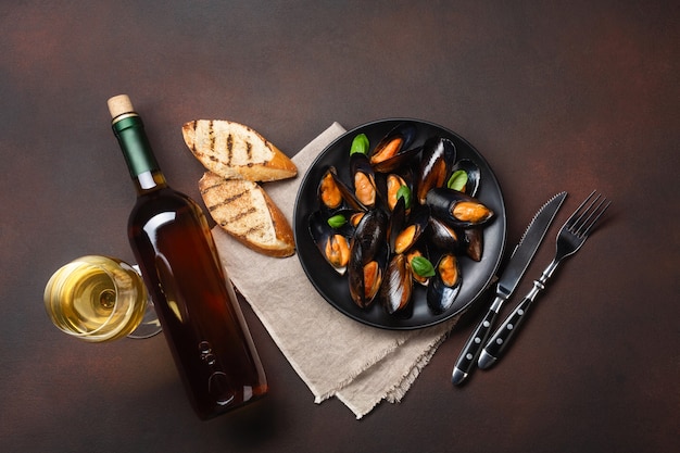 Moules de fruits de mer et feuilles de basilic dans une assiette noire avec bouteille de vin, verre à vin, tranches de pain, fourchette, couteau, toile de jute sur fond rouillé. Vue de dessus.