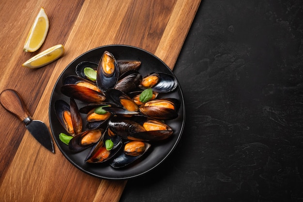 Moules de fruits de mer et feuilles de basilic dans une assiette noire avec citron, couteau sur planche de bois et table en pierre. Vue de dessus avec place pour votre texte.