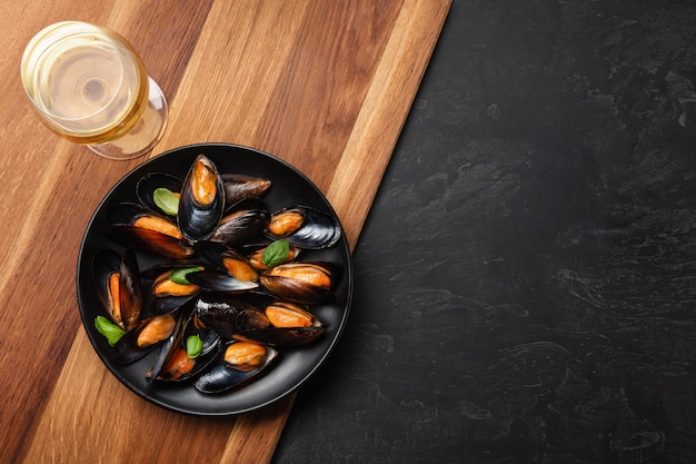 Moules de fruits de mer et feuilles de basilic dans une assiette noire avec verre à vin, citron, couteau sur planche de bois et table en pierre