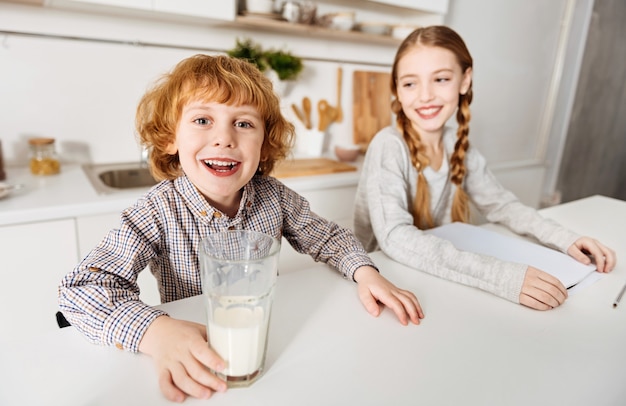 Moustache laiteuse. Joyeux et charmants frères et sœurs adorables commençant leur journée ensemble pendant que le garçon buvant du lait et sa sœur font son devoir à la maison