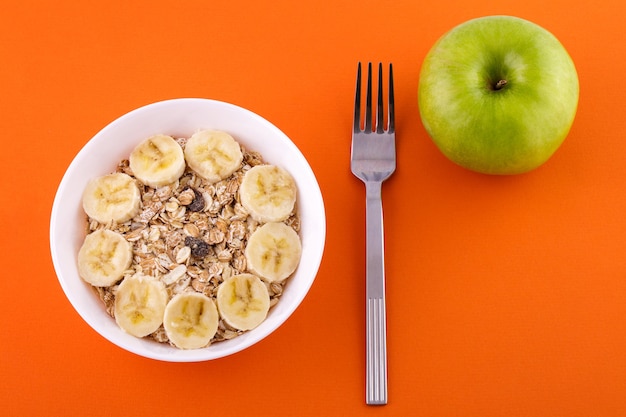 Muesli à la banane dans une assiette blanche sur fond orange une fourchette et une pomme verte aliment sain a...