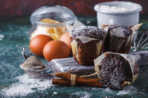 Muffin au chocolat et aux carottes saupoudré de sucre glace, d’une tasse de thé et d’ingrédients de cuisson. Farine, oeufs, citron citrus sur une table sombre