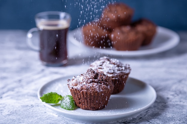 Muffins au chocolat à la menthe sur une plaque noire, parsemés de sucre glace. Cuisson maison. En arrière-plan, une tasse de café et une assiette avec des muffins. Table en marbre et fond bleu.