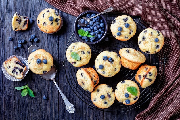 Muffins aux myrtilles cuits avec des myrtilles fraîches sur une grille de refroidissement noire avec de la menthe fraîche sur le dessus sur une surface en bois rustique sombre, vue de dessus, gros plan