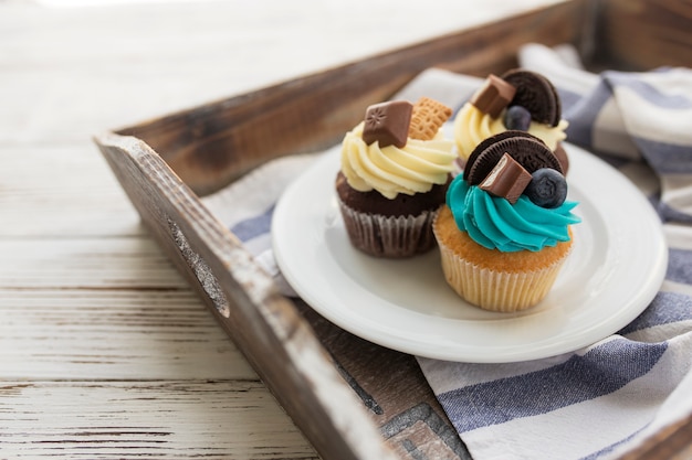 Muffins et cupcakes multicolores savoureux sur la plaque blanche sur un plateau en bois avec une serviette. Décoré avec différents bonbons, biscuits et fromage à la crème sucré coloré sur le dessus.