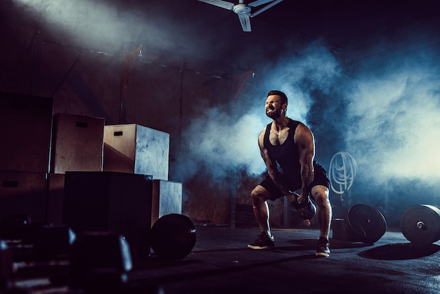 Musclé attrayant caucasien homme tatoué barbu soulevant kettlebell dans une salle de sport