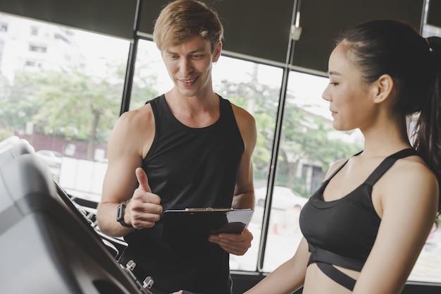 Photo musculaire beau caucasien entraîneur personnel homme montrant les pouces vers le haut avec jeune femme asiatique dans la salle de fitness