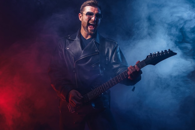 Un musicien de heavy metal barbu brutal en veste de cuir et lunettes de soleil joue de la guitare électrique sur du noir avec de la fumée