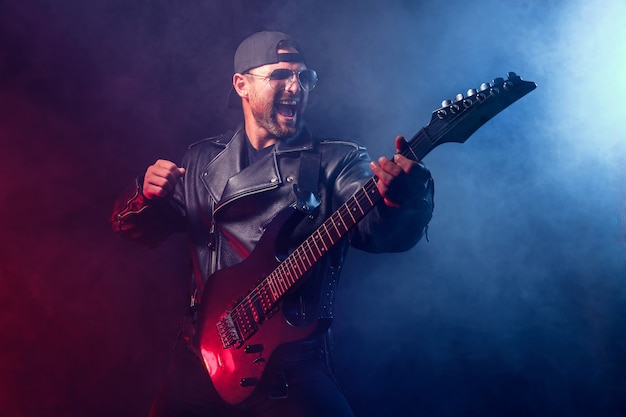 Un musicien de heavy metal barbu brutal en veste de cuir et lunettes de soleil joue de la guitare électrique sur fond noir