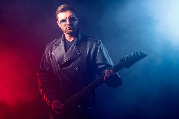 Un musicien de heavy metal barbu brutal en veste de cuir et lunettes de soleil joue de la guitare électrique sur fond noir