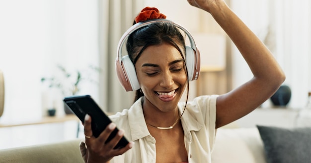 Photo la musique du smartphone et la danse d'une femme heureuse sur le canapé dans la célébration à la maison et l'écoute d'une application audio téléphone personne indienne excitée sur les écouteurs et le son de la radio ou la liberté dans le salon sur la technologie mobile