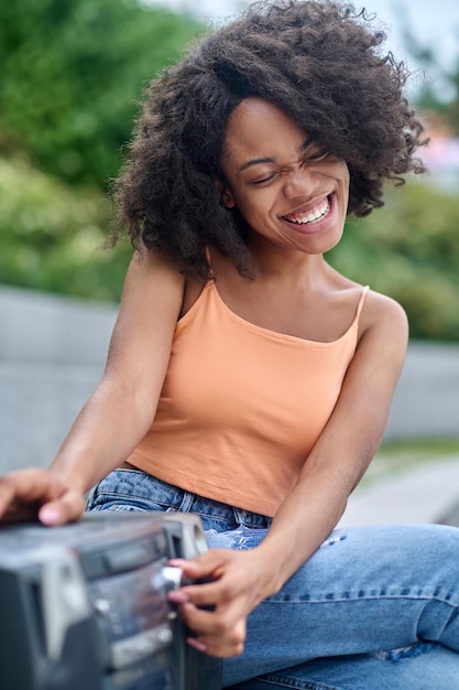 Musique, plaisir. Souriante jolie jeune femme à la peau foncée avec les yeux fermés et les cheveux bouclés touchant le plateau tournant en écoutant de la musique assis dans le parc par beau temps