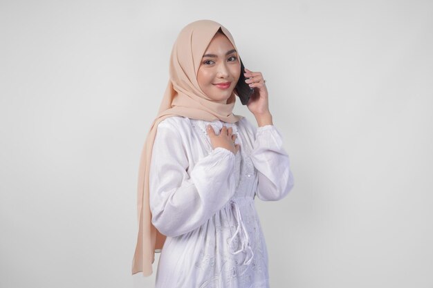 Photo une musulmane asiatique heureuse portant une robe blanche et un hijab tenant son smartphone et faisant un appel à sa famille ou à un ami isolée par un fond blanc concept de ramadan et eid mubarak
