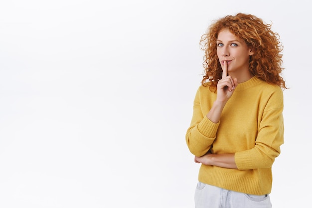 Mystérieuse et impertinente belle femme rousse séduisante aux cheveux bouclés en pull jaune, préparez un incroyable cadeau surprise pour son petit ami, taisez-vous avec l'index pressé sur les lèvres, souriant sournoisement