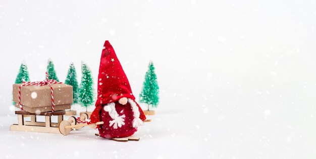 Le nain scandinave Nisse en vêtements rouges avec des skis dans les mains avec des nattes qui sortent Le gnome porte un traîneau avec un cadeauIl y a des arbres de Noël en arrière-plan