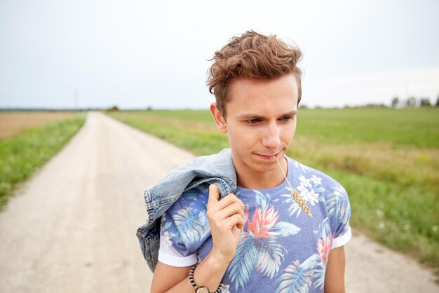 nature, été, culture des jeunes et concept de personnes - triste jeune hippie marchant le long d'une route de campagne mâchant un pic de seigle