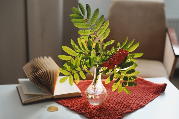 Nature morte d'automne avec une branche de sorbier dans un vase et un livre et des pommes dans un intérieur confortable