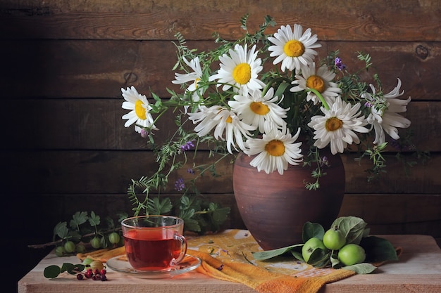 Nature morte estivale avec un bouquet de pâquerettes, de pommes vertes, un brin de groseilles à maquereau et un verre de compote.