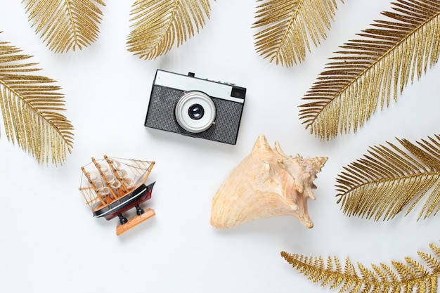 Nature morte de voyage minimaliste. Coquillages, appareil photo rétro parmi les feuilles de palmier dorées décoratives sur fond blanc. Vue de dessus