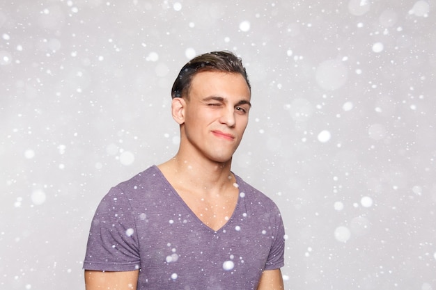 Neige, hiver, Noël, émotion, publicité et concept de personnes - Beau jeune homme sur fond de neige en regardant la caméra. Portrait d'un jeune homme riant dans les poches appuyé contre un mur gris.