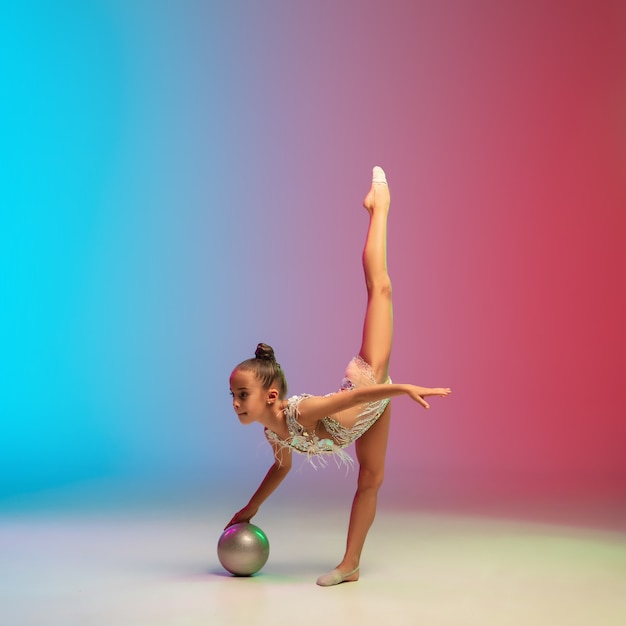 Énergie. Petite fille caucasienne, entraînement de gymnaste rythmique, effectuant isolée sur fond de studio dégradé bleu-rouge en néon. Enfant gracieux et flexible, fort. Concept de sport, mouvement, action.