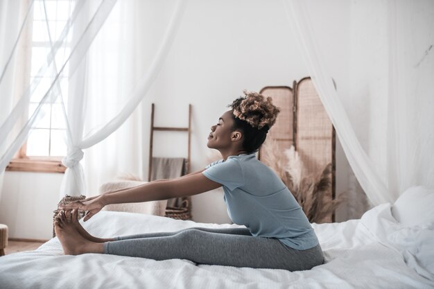 Énergie positive. Jeune femme afro-américaine satisfaite dans des vêtements à la maison assis sur le lit tenant ses orteils en méditant