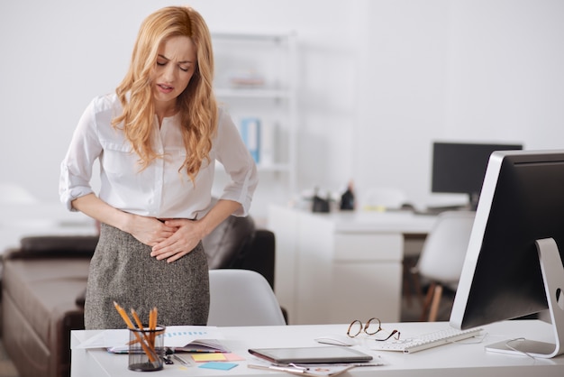 Nerveuse jeune femme d'affaires professionnelle debout dans le bureau et travaillant tout en touchant son ventre et souffrant de douleurs