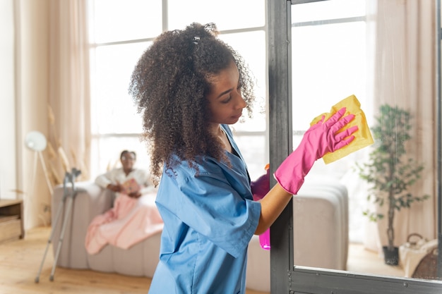 Nettoyage de l'aidant. Soignant afro-américain bouclé nettoyant la porte en verre à la maison du patient