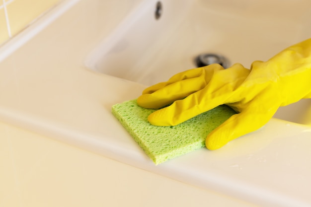 Photo nettoyage du lavabo et du robinet de salle de bain avec un détergent dans des gants en caoutchouc jaune et une éponge verte