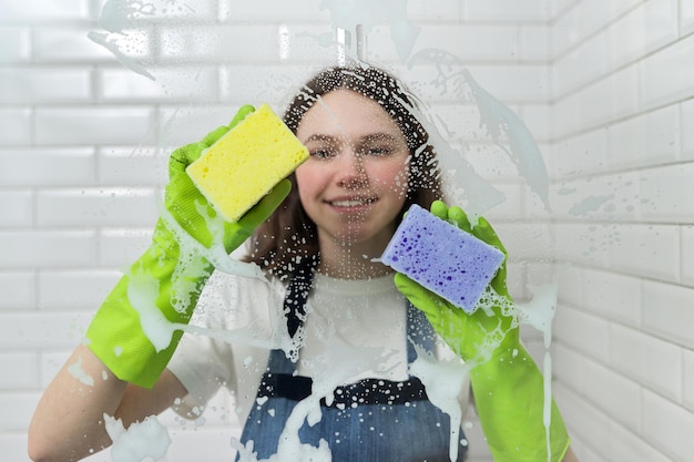 Nettoyage de la salle de bain, adolescente lavant le verre de la douche avec de la mousse et des éponges, gros plan de la main des femmes dans des gants verts et des éponges colorées