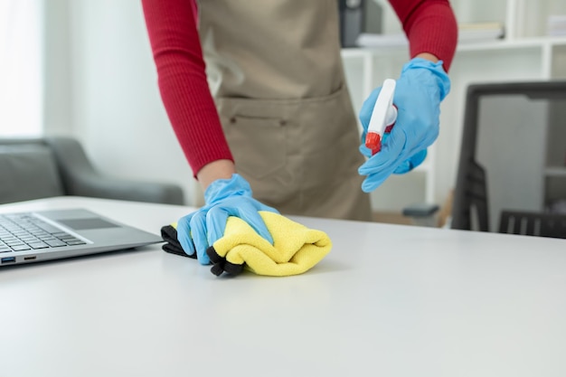 Photo nettoyer la surface du bureau au bureau avec un spray désinfectant porter des gants et essuyer la table avec une serviette la femme de ménage nettoie le bureau de travail pour l'hygiène à cause de l'idée de nettoyage covid19