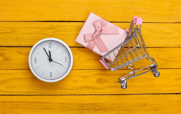 Noël, achats du nouvel an. Chariot de supermarché mini avec boîte-cadeau et horloge sur une surface en bois jaune. Vue de dessus