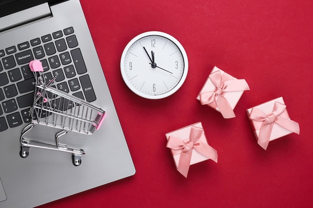 Photo noël, achats en ligne du nouvel an. ordinateur portable, mini chariot de supermarché avec coffrets cadeaux, horloge sur fond rouge. vue de dessus