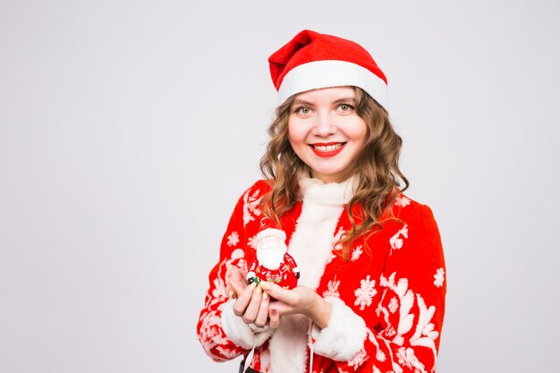 Noël, célébration, concept de personnes - femme drôle en costume de Santa tenant une figurine de Santa sur un mur blanc