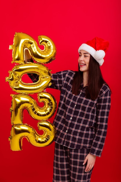 Photo noël , concept de joyeuses fêtes - une jeune femme en pyjama et un chapeau de père noël détient les numéros 2022