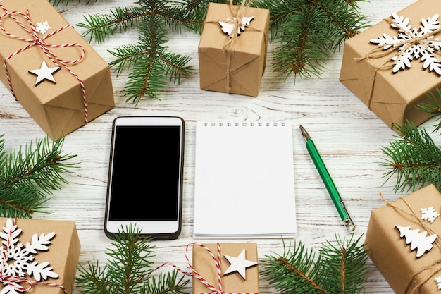 Noël à faire la liste. Smartphone, bloc-notes, coffret cadeau avec ruban rouge, liste de courses avec espace de copie sur table en bois blanc. Concept de vacances d'hiver. Vue de dessus.