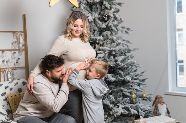 Noël Famille Bonheur Portrait de papa maman enceinte et petit fils assis fauteuil à la maison près de l'arbre de Noël câlin sourire européen jeune adulte vacances en famille matin