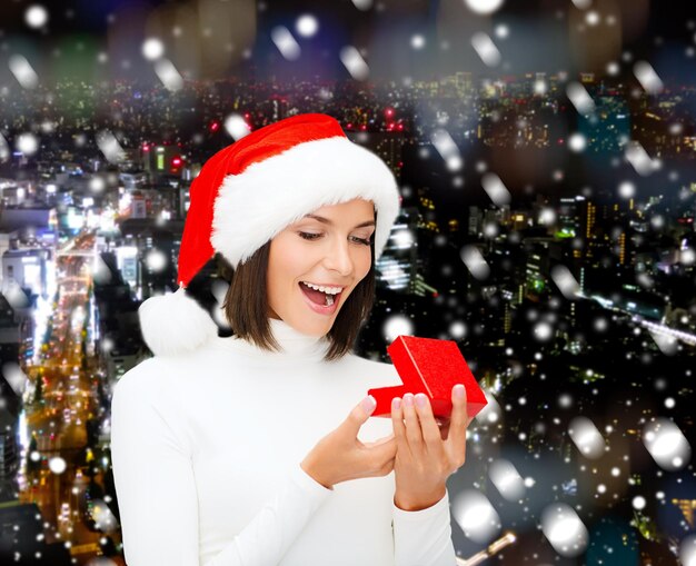 Noël, hiver, bonheur, vacances et concept de personnes - femme souriante en bonnet d'assistance avec boîte-cadeau sur fond de ville de nuit enneigée