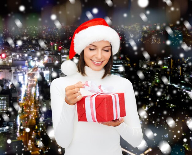 Noël, hiver, bonheur, vacances et concept de personnes - femme souriante en bonnet d'assistance avec boîte-cadeau sur fond de ville de nuit enneigée