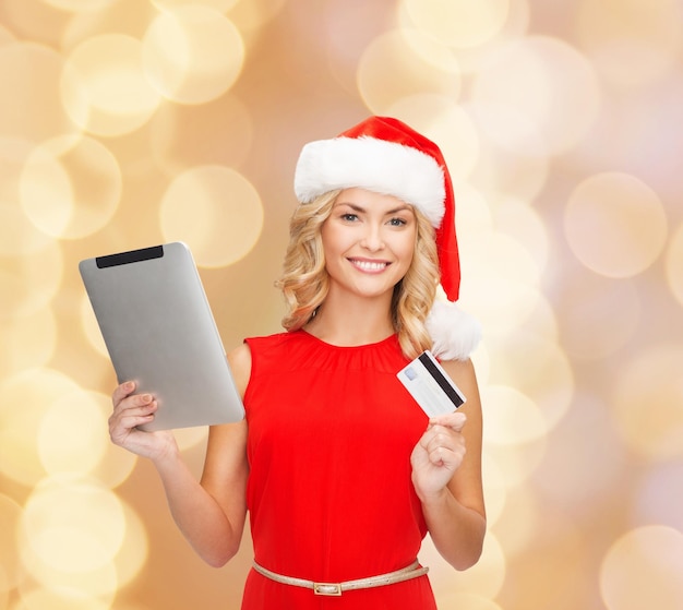 Noël, technologie, shopping et concept de personnes - femme souriante en bonnet d'assistance avec ordinateur tablette et carte de crédit sur fond de lumières beiges