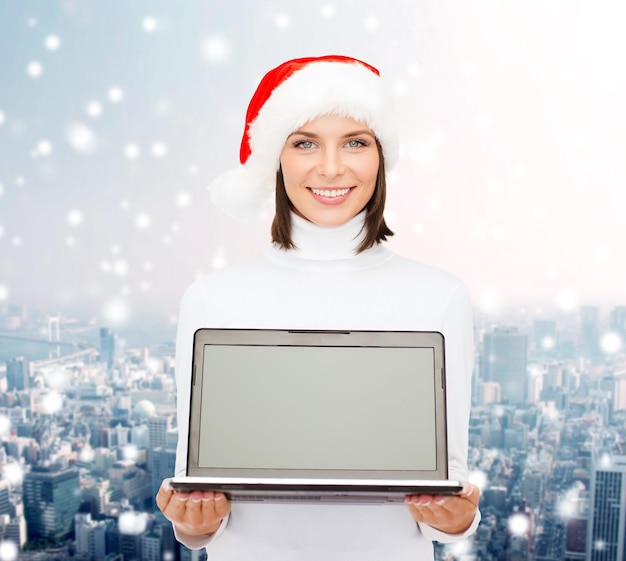 Noël, technologie, vacances d'hiver et concept de personnes - femme souriante en bonnet d'assistance avec ordinateur portable à écran blanc sur fond de ville enneigée