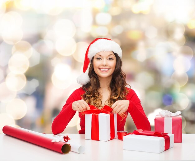 noël, vacances, célébration, décoration et concept de personnes - femme souriante en bonnet d'assistance avec papier de décoration emballage coffrets cadeaux sur fond de lumières