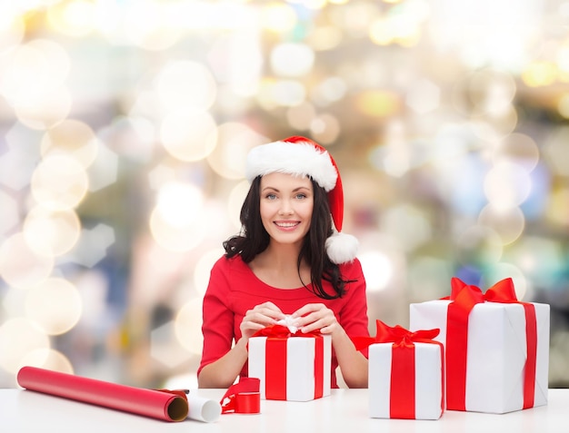 noël, vacances, célébration, décoration et concept de personnes - femme souriante en chapeaux de santa helper avec papier de décoration et coffrets cadeaux sur fond de lumières