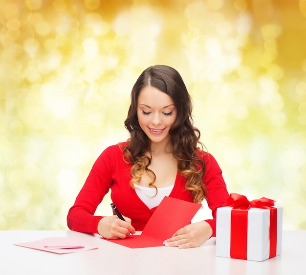 noël, vacances, célébration, salutation et concept de personnes - femme souriante avec boîte-cadeau écrivant une lettre ou envoyant une carte postale sur fond de feux jaunes