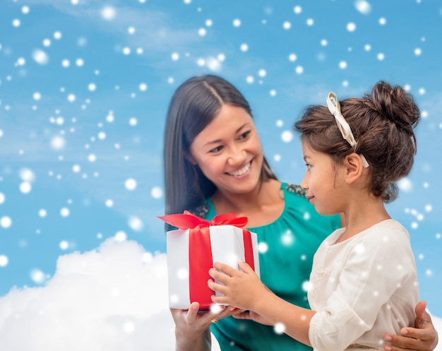 Noël, vacances, concept de famille et de personnes - heureuse mère et enfant fille avec boîte-cadeau sur ciel bleu enneigé avec fond nuageux