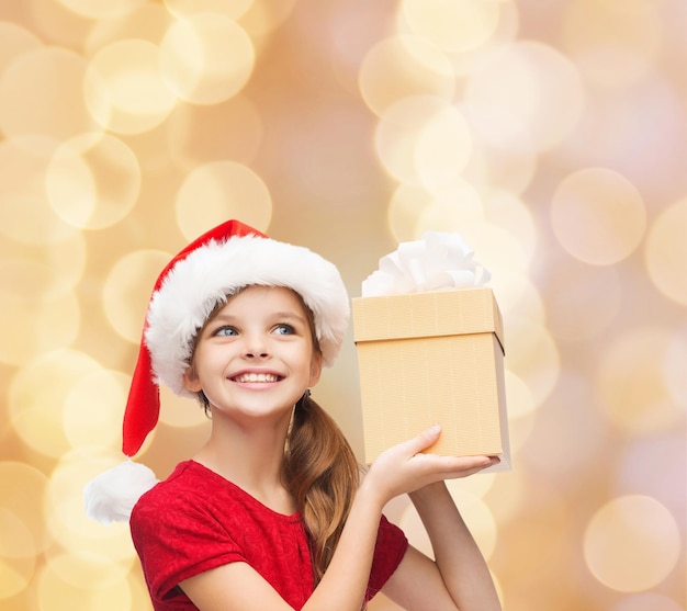 Noël, vacances, enfance et concept de personnes - jeune fille souriante en bonnet d'assistance avec boîte-cadeau sur fond de lumières beiges