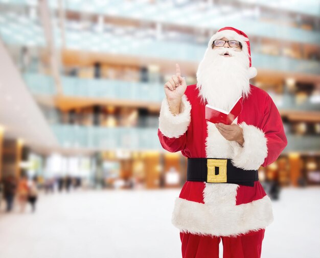 noël, vacances, geste et concept de personnes - homme en costume de père noël avec bloc-notes pointant le doigt vers le haut sur fond de centre commercial