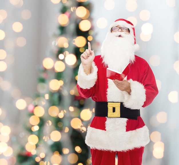 noël, vacances, geste et concept de personnes - homme en costume de père noël avec bloc-notes pointant le doigt vers le haut sur fond de lumières d'arbre