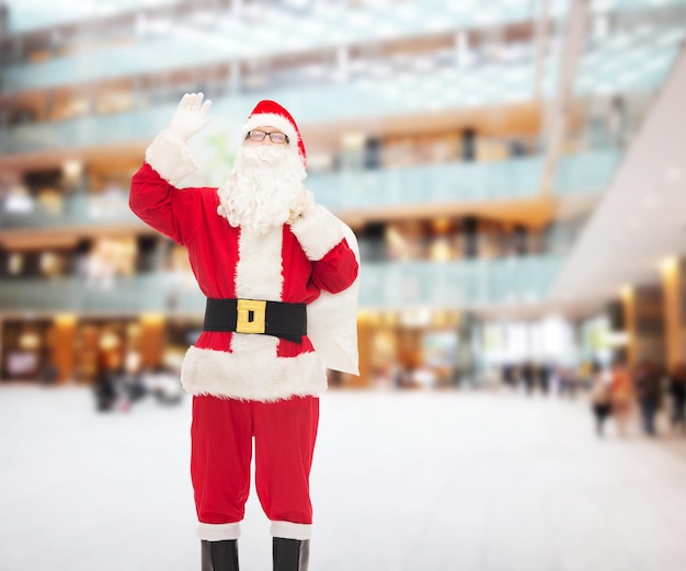 noël, vacances, geste et concept de personnes - homme en costume de père noël avec sac agitant la main sur fond de centre commercial