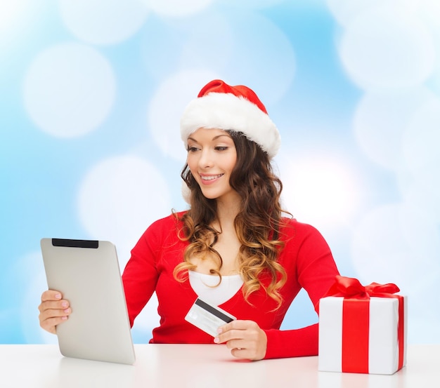 Noël, vacances, technologie et concept de shopping - femme souriante en bonnet d'assistance avec boîte-cadeau, carte de crédit et ordinateur tablette sur fond de lumières bleues